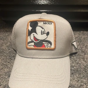 Grå keps med Mickey Mouse patch - Snygg grå keps med broderad Mickey Mouse-patch framtill och mesh på sidorna för extra ventilation. Kepsen har justerbar snapback och en liten Mickey-detalj på sidan. Perfekt för dig som gillar Disney och vill ha en cool accessoar.