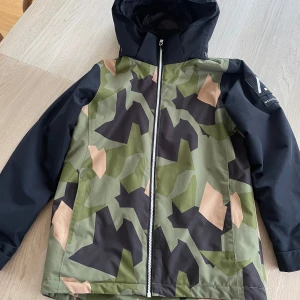 Camouflage vindjacka från WearColour - Skidjacka/vinterjacka från WearColour i storlek 160 med grön, svart och beige camouflage-mönster. Jackan har svart huva och ärmar, hel dragkedja framtill och logga på ärmen. Perfekt för dig som gillar streetstyle. 