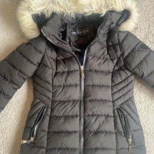 Svart dunjacka med fuskpäls från Polo Club - Snygg svart dunjacka från Polo Club med fluffig fuskpäls på huvan. Jackan har quiltad design, dragkedjor på fickorna och framtill. Perfekt för kalla dagar.