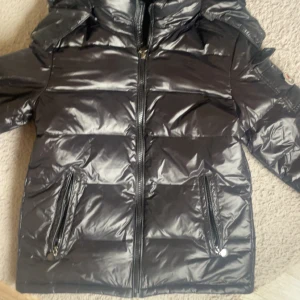 Svart pufferjacka  - Snygg svart pufferjacka från med glansig finish och avtagbart märke på ärmen. Jackan har dragkedja framtill, två fickor med dragkedja och hög krage. Perfekt för kalla dagar när du vill hålla stilen och värmen. Denna efterliknar moncler men är inte den.