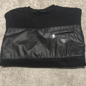 Prada Sweatshirt - Svart långärmad sweatshirt från Prada med unik skinnliknande panel över bröstet, liten ficka med dragkedja och ikonisk Prada-logga. Tillverkad i bomull, perfekt för dig som gillar stilrena och exklusiva plagg med coola detaljer.