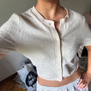 Beige kortärmad kofta i stickat - Superfin beige kofta i stickat material med korta ärmar och rund hals. Modellen är croppad och har ribbade detaljer vid hals och knappslå. Perfekt att slänga över ett linne eller klänning för en chill look.