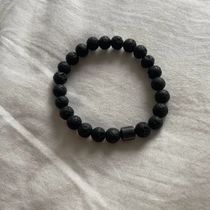 Svart armband med lavapärlor - Snyggt svart armband med runda lavapärlor och en större cylinderformad pärla som detalj. Armbandet har en cool, lite rå känsla tack vare de porösa lavastenarna och passar perfekt för dig som gillar minimalistiska accessoarer.