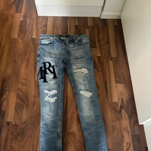 Distressed ljusblå jeans från Amiri - Säljer ett par ljusblå jeans från Amiri med slitna detaljer och patchade hål. Jeansen har stora svarta bokstavsbroderier på låret och baksidan, klassisk femficksmodell och skinny fit. Materialet är 98% bomull och 2% elastan för extra stretch.