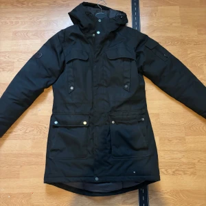 Svart parkajacka från Everest - Svart parkajacka från Everest med huva och flera praktiska fickor framtill. Jackan har långa ärmar, dragkedja och tryckknappar. Perfekt för kalla dagar och har en stilren, enkel design som funkar till det mesta.