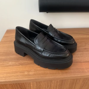 Chunky svarta loafers i skinn - Säljer ett par svarta loafers med chunky platt sula och klassisk pennyloafers-design. Skorna är i skinn och har en rund tå samt grov yttersula. Är små i storleken 37 (36).