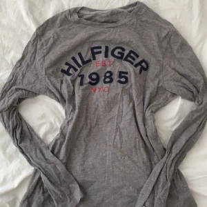 Grå långärmad tröja Tommy Hilfiger - Snygg grå långärmad tröja från Tommy Hilfiger med tryck 'HILFIGER 1985 NYC' i marinblått och rött framtill. Mjuk och tunn i bomull, perfekt till jeans eller kjol. Klassisk rund halsringning och relaxed fit.