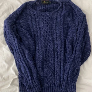 Blå stickad tröja från John Howard - Mysig blå stickad tröja från John Howard med grovt kabelstickat mönster och rund hals. Tröjan har långa ärmar och breda ribbade muddar vid ärmslut och nederkant. Perfekt för lager-på-lager och chill höst- och vinterstil.