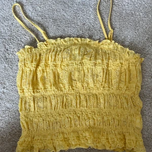 Gul broderad topp från BikBok - Supersöt gul topp från BikBok med smala axelband och broderade detaljer. Toppen är smockad och har volangkant upptill och nertill. Perfekt för soliga dagar och ger en riktigt somrig vibe! 
