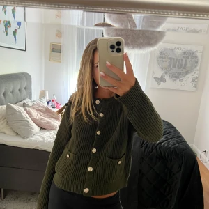 Mörkgrön stickad kofta från H&M - Mysig mörkgrön stickad kofta från H&M med snygga guldiga knappar framtill och två fickor. Koftan har långärmad passform och grovstickad struktur som ger en cool och avslappnad vibe. Perfekt att slänga över en t-shirt eller linne.