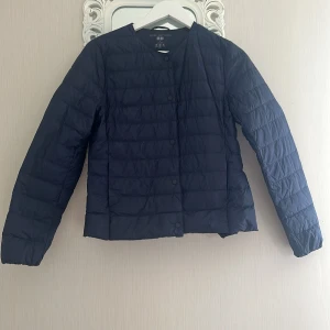 Mörkblå lättviktsdunjacka från Uniqlo S - Säljer en mörkblå ultralätt dunjacka från Uniqlo med rund halsringning och tryckknappar framtill. Jackan är quiltad och har en clean, minimalistisk look som passar perfekt till lager-på-lager-stil. Långärmad och superenkel att bära med sig.