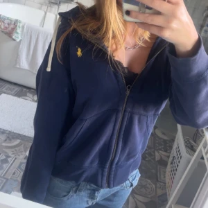 blå hoodie från Polo Ralph Lauren - Säljer en marinblå hoodie från Polo Ralph Lauren med dragkedja och huva. Tröjan har vit snörning i huvan, klassisk gul logga på bröstet och är i mjuk bomull. Perfekt för chill dagar och snygg till jeans.
