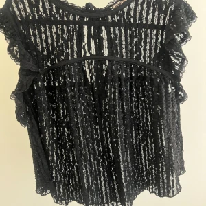 Svart spetsblus med volangärmar - Supersnygg svart blus i spets med tunna vertikala ränder och volangdetaljer på ärmarna. Blusen är ärmlös med knytning i nacken och har en transparent look som ger en cool vibe. Perfekt att styla med jeans eller kjol för en trendig outfit. Har aldrig använt!💗