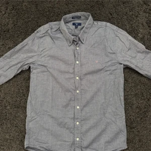 Grå oxfordskjorta från Gant - Klassisk grå oxfordskjorta från Gant med button down-krage och vita knappar. Skjortan har lång ärm och en liten broderad Gant-logga på bröstet. Tillverkad i bomull och har en tidlös, normal passform som funkar till det mesta.