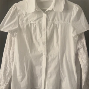 Vit kortärmad skjorta med krage - Snygg vit skjorta med korta ärmar och klassisk krage. Skjortan har knappar framtill och en lätt rynkad detalj vid axlarna. Perfekt för en clean och stilren look. Tillverkad i bomull för en fräsch känsla.