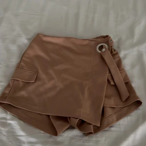 Beige omlottshorts från Kechi Woman - Snygga beige shorts från Kechi Woman i omlottmodell med bred midja och coolt bältdetalj med metallring. Shortsen har fickor på sidorna och är tillverkade i ett mjukt, slätt material som ger en clean och trendig look.
