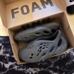 Adidas Yeezy Foam Runner svart - Säljer ett par Adidas Yeezy Foam Runner i svart. Skorna har en futuristisk design med många hål för ventilation och är tillverkade i lätt syntetmaterial. Perfekta för dig som vill sticka ut med en unik och bekväm look.