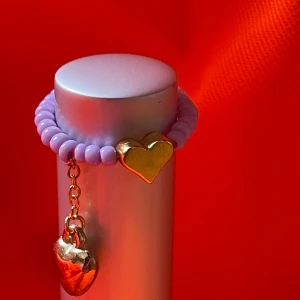 By prisförslag- Lila pärlring med guldhjärtan - Supergullig ring med lila pärlor och två guldiga hjärtan – ett fast och ett som hänger i kedja. Perfekt accessoar för att ge din outfit en söt och trendig touch. Stretchig modell 