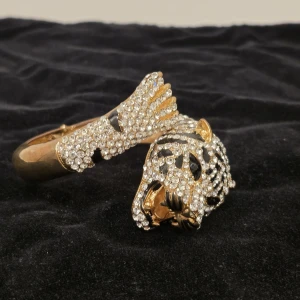 Glittrigt guldarmband med tigerhuvud - Unikt armband i guldton med ett detaljerat tigerhuvud och tass, täckt av gnistrande stenar. Svarta detaljer skapar ett coolt mönster som påminner om en leopard eller tiger. Perfekt statement piece för dig som vill sticka ut.