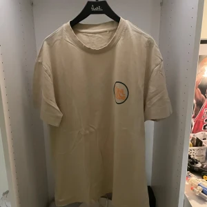 Beige t-shirt från Primark med tryck - Säljer en beige t-shirt från Primark i storlek S. T-shirten har ett litet tryck på bröstet med texten 'It's a Vibe' i orange och vitt, inramat av en svart cirkel. Klassisk passform och rund hals. Perfekt för chill dagar. Knappt använd