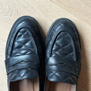 Svarta quiltade loafers i skinn från Flattered - Snygga svarta loafers från Flattered med quiltad design och chunky sula. Skorna är tillverkade i skinn och har klassisk pennyloafers-detalj framtill. Perfekta för dig som vill ha en stilren men edgy look till vardags. Passar lika bra till jeans som till kostymbyxor.