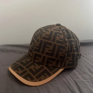 Fendi keps  - Snygg keps från Fendi med ikoniskt FF-monogram i svart och brunt över hela kepsen. Justerbar rem baktill i brunt läder och böjd skärm med beige kant. Tillverkad i Italien, perfekt för dig som vill sticka ut med lyxig streetstyle.
