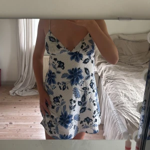 Vit och blå blommig kortklänning från Gina  - Supersöt kortklänning från ginatricot med tunna axelband och v-ringning. Klänningen är vit med stora blå blommor och har en lätt strukturerad yta. .