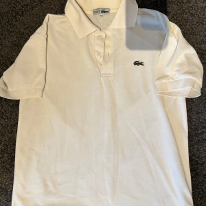 Lacoste piké  - Klassisk vit Lacoste piké i mycket fint skick! Tidlös design med det ikoniska krokodilmärket på bröstet. Perfekt för både vardag och lite mer uppklädda tillfällen. Skön passform och hög kvalitet i bomull – ett måste i garderoben! Perfekt för sommaren, semestern eller som en stilren bas i din outfit.