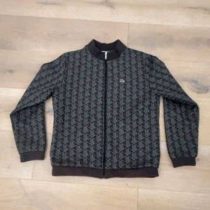 Svart kofta från Lacoste - Snygg svart kofta från Lacoste med grått geometriskt mönster och klassisk krokodillogga på bröstet. Koftan har ribbade muddar, dragkedja framtill och en cool, sportig vibe. Storlek 164cm (XS), den är köpt från kidsbrandstore för 1499kr, använd väldigt lite är som ny 9/10 skick! perfekt för dig som vill sticka ut med något stilrent ✅