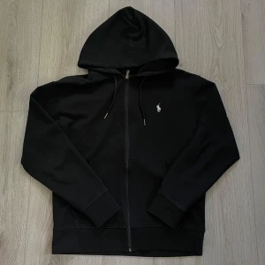 Svart hoodie från Polo Ralph Lauren - Svart hoodie från Polo Ralph Lauren med dragkedja och huva. Klassisk design med vit broderad logga på bröstet. Skön passform och ribbade muddar. Storlek M. 9/10, hör av er vid frågor