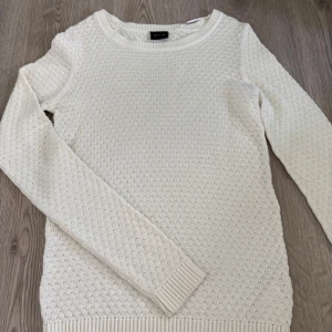 Vit stickad tröja från Vila - Snygg vit stickad tröja från Vila med diskret strukturmönster. Tröjan har rund halsringning, långa ärmar och ribbade muddar vid ärmslut och nederkant. 