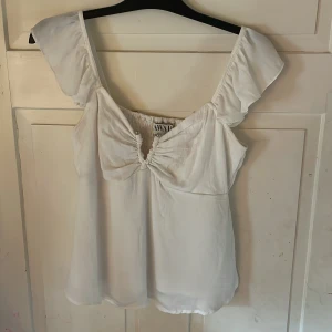 Vit blus med volangärm från AWARE - Supersöt vit blus från AWARE med volangärmar och smockad rygg. Toppen har en snygg rynkad detalj framtill med små silvriga knappar och sitter lite tajt vid bröstet sedan luftigare nedtill. Använd en gång. 