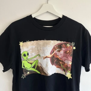 Svart t-shirt med alien-tryck - Svart t-shirt med en kreativ twist på Michelangelos Skapelsen. T-shirten har rund hals och är i mjuk bomull. Köpt i New York. 