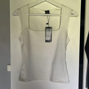 Vit tanktopp från vero Moda - Supersnygg vit linnetopp från vero moda med breda axelband och fyrkantig ringning. Toppen är ärmlös och har en clean, stilren look som passar perfekt till jeans eller kjol. Materialet är mjukt och stretchigt för en bekväm passform💓🌟