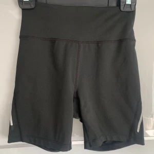 Svarta högmidjade träningsshorts SOC - Svarta högmidjade träningsshorts från SOC med tight passform. Tillverkade i stretchigt syntetmaterial, perfekta för löpning eller gym. Reflekterande detalj på benet för extra synlighet. Med en ficka för mobil eller annat i ryggen. 