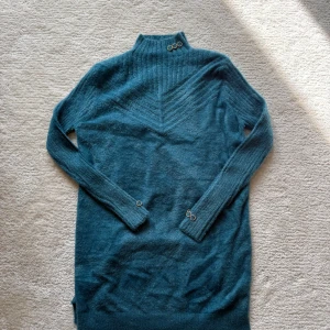 Mörkblå stickad långtröja - Mysig mörkblå stickad långtröja med polokrage och dekorativa metallringar vid halsen och ärmsluten. Klänningen har långa ärmar och ribbade detaljer upptill som ger en snygg struktur. Perfekt för kalla dagar när du vill vara både bekväm och snygg.