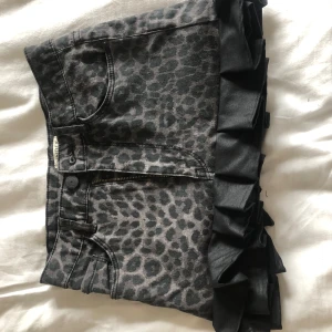 Grå leopardmönstrad jeanskjol med volang - Cool grå jeanskjol med leopardmönster och svarta volanger längst ner. Kjolen har fickor både fram och bak samt dragkedja och knapp framtill. Perfekt för dig som vill sticka ut lite extra.