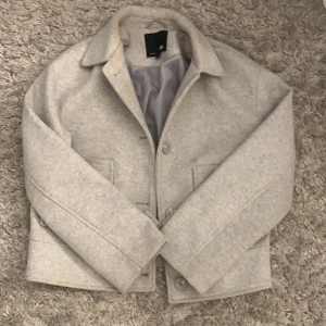 Ljusgrå ulljacka från Lager 157 - Snygg ljusgrå jacka i ullblandning från Lager 157 med klassisk krage och knäppning framtill. Jackan har två fickor och är helfodrad för extra komfort. Perfekt för kyligare dagar.