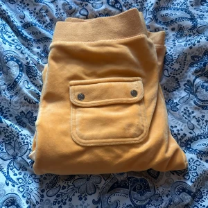 Orange mjukisbyxor från Juicy Couture - Säljer ett par mjukisbyxor från Juicy Couture i en snygg orange färg. Byxorna har resår och snörning i midjan samt en ficka med knappar bak. Perfekta för en avslappnad stil.