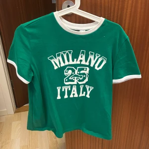 Grön t-shirt med Milano-tryck - Grön t-shirt med vita detaljer och texten 'Milano 25 Italy' framtill. T-shirten har rund halsringning och korta ärmar med vita kanter. Perfekt för en sportig och avslappnad stil.