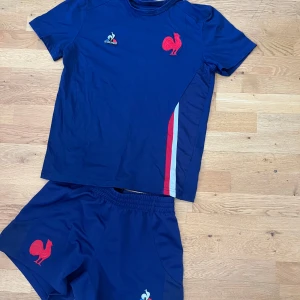 Blå Frankrike fotbollströja från Le Coq Sportif - Säljer en blå fotbollströja från Le Coq Sportif med röd tupp-logga och franska flaggans färger på sidan