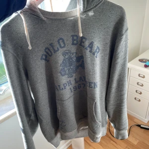 Grå hoodie från Ralph Lauren Polo Bear - Säljer en grå hoodie från Ralph Lauren med Polo Bear-tryck framtill och texten '1967'. Tröjan har huva med dragsko och känguruficka. Har inte använt tröjan jätte mycket, köpte den förra sommaren.