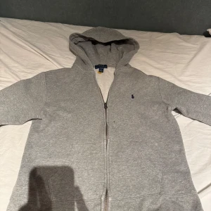 Grå hoodie från Polo Ralph Lauren - Säljer en klassisk grå hoodie från Polo Ralph Lauren med dragkedja och huva. Tröjan har ett litet broderat logomärke på bröstet och är fodrad med mjukt material på insidan. Perfekt för en avslappnad stil. Storleken skulle jag beskriva som mindre för den har krympt i tvätten och är ungefär för 158cm långa 