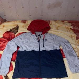 Vindjacka med röd huva från Jack & Jones - Snygg vindjacka från Jack & Jones med röd huva, ljusgrå överdel och mörkblå nederdel. Jackan har dragkedja framtill och en bröstficka med dragkedja. Perfekt för blåsiga dagar.