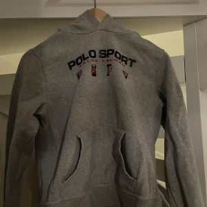 Grå hoodie från Polo Sport Ralph Lauren - Säljer en grå hoodie från Polo Sport Ralph Lauren med tryckt logga och emblem på bröstet. Tröjan har huva och en stor magficka framtill. Perfekt för en sportig och avslappnad stil. Säljer: pga för liten. Bra skick. JUNIOR LARGE. Pris kan diskuteras.