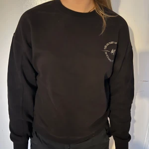 Svart sweatshirt med tryck - Svart sweatshirt med rund hals och diskret vitt tryck på bröstet. Tröjan har en klassisk passform och långa ärmar. Perfekt för en avslappnad stil.