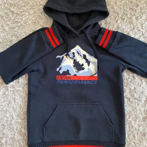 Svart hoodie från Peak Performance med röda detaljer - Svart hoodie från Peak Performance med långa ärmar och röda samt svarta ränder på ärmslut och nederkant. Stor logga med bergsmotiv framtill och känguruficka. Perfekt för en sportig och avslappnad stil. Supermysig hoddie som är sparsamt använd.