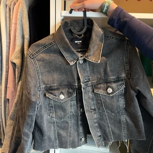Svart croppad jeansjacka med mönster - Säljer en svart croppad jeansjacka från Bik Bok med silvriga knappar och två bröstfickor. Jackan har ett unikt cirkelmönster på ryggen och rå, fransig nederkant. Perfekt för dig som vill sticka ut med en cool och personlig stil.