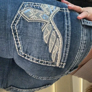 Blå jeansbyxor med broderade bakfickor - Säljer ett par blå jeansbyxor med snygga broderade detaljer på bakfickorna. Byxorna är lågmidjade och har bootcut. Helt nya utan prislapp