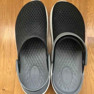 Svarta Crocs LiteRide clogs - Säljer ett par svarta Crocs LiteRide clogs med grå insida och vit sula. Skorna har perforerat mönster för ventilation och justerbar hälrem. Perfekta för avslappnade dagar och enkel att ta på och av.